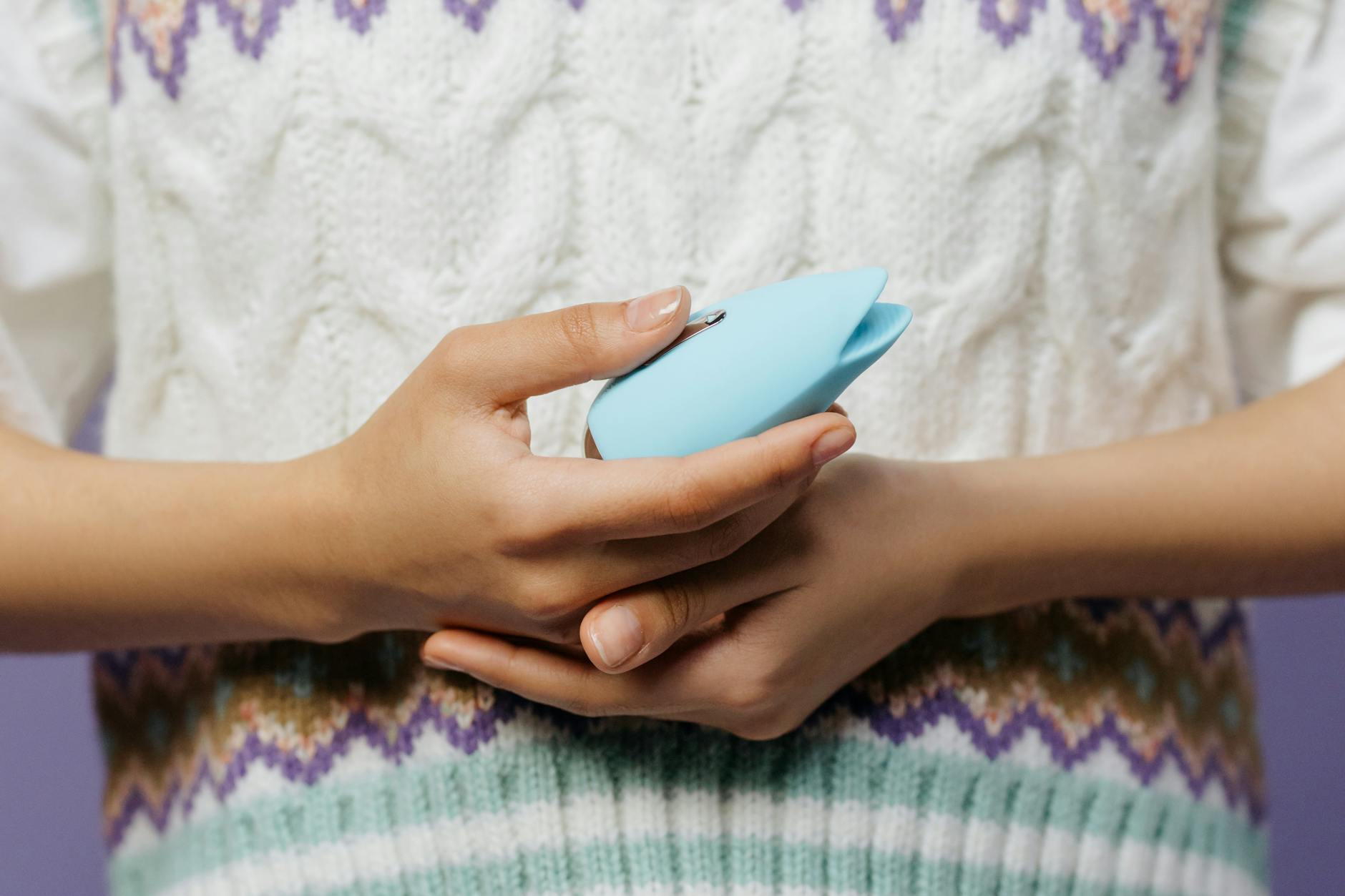 Close-up of hands holding a blue personal massager against a knitted sweater, representing postpartum pleasure recovery.